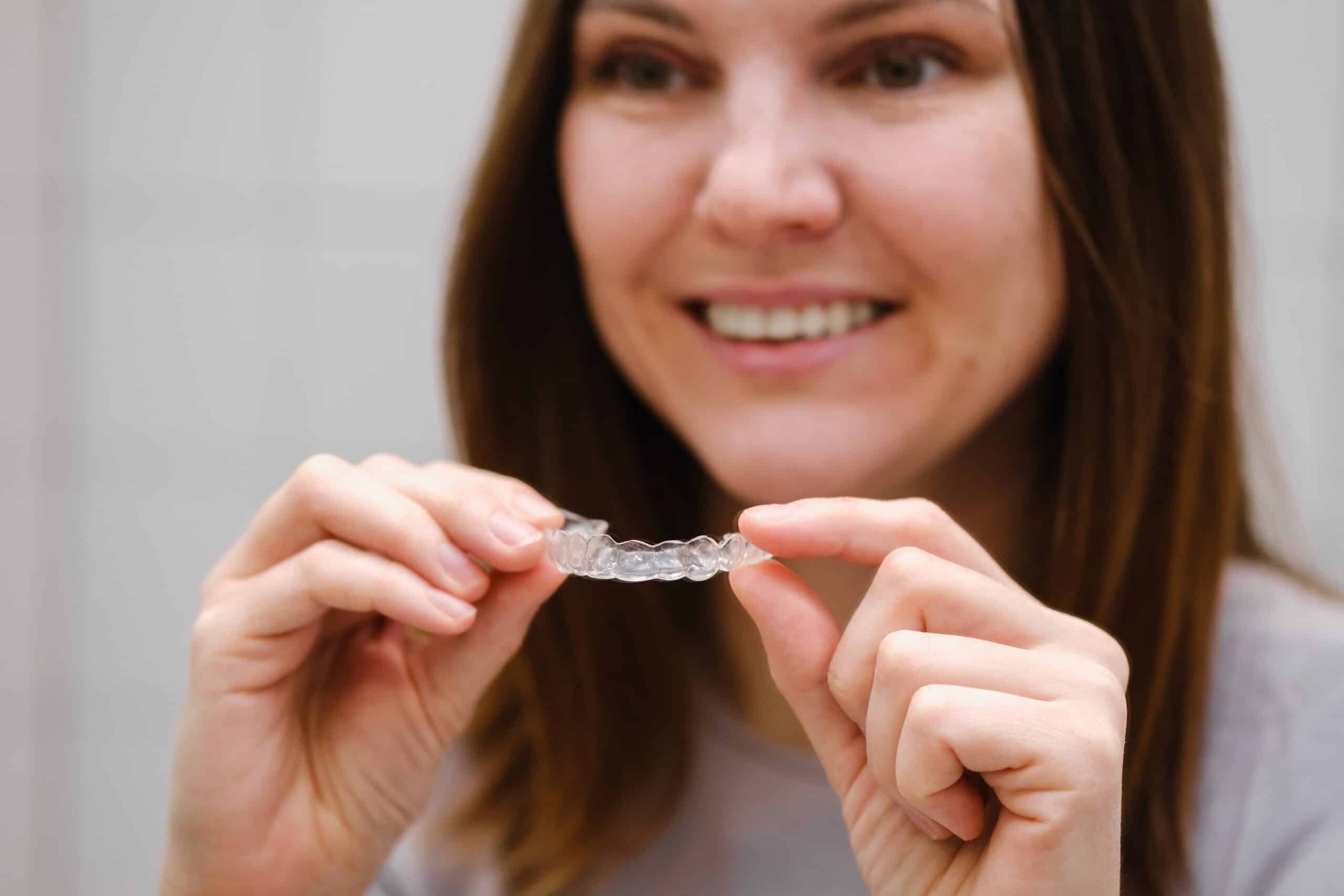 A smiling woman holds a clear dental aligner with both hands. The focus is on the aligner, highlighting its transparency and detailed ridges.